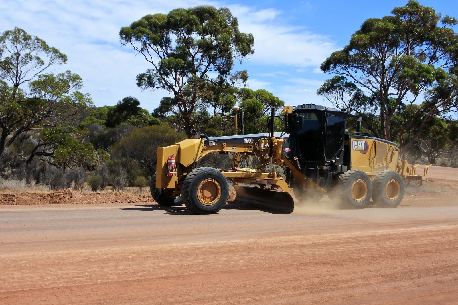 A motor grader working the formation of an unsealed road through eucalypt scrub in regional Western Australia.